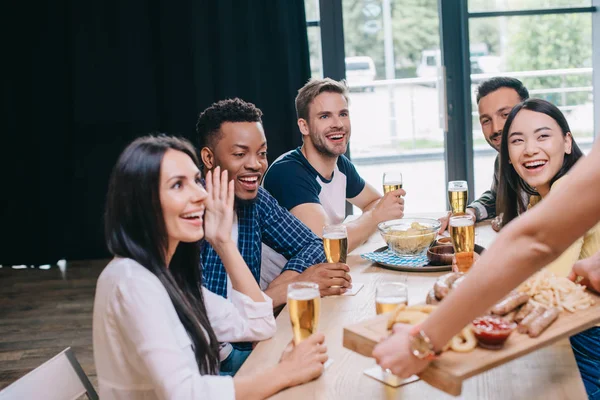 Cropped view of woman holding tray with snacks near happy multicultural friends in bar — Stock Photo