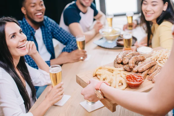 Cropped view of woman holding tray with fried sausages, onion rings and french fries near happy multicultural friends in bar — Stock Photo