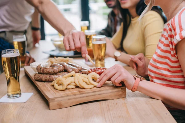 Vista parcial de amigos multiculturales sentados en el pub cerca de la bandeja con aros de cebolla frita, salchichas y papas fritas - foto de stock