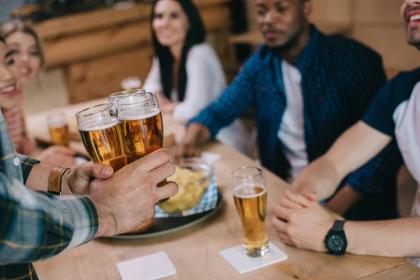 Vista recortada del hombre sosteniendo vasos de cerveza ligera cerca de amigos multiculturales en el pub - foto de stock