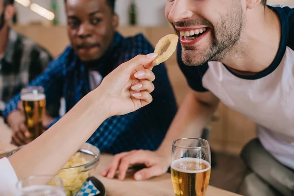 Vista recortada de la mujer alimentando hombre alegre con anillo de cebolla frita mientras está sentado en el pub con amigos - foto de stock