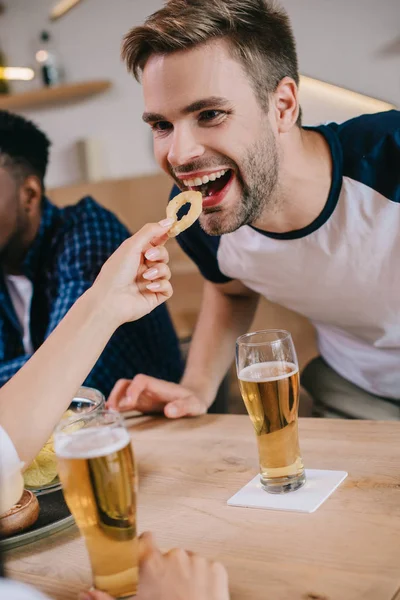 Partial view of woman feeding cheerful man with fried onion ring while sitting in pub with multicultural friends — Stock Photo