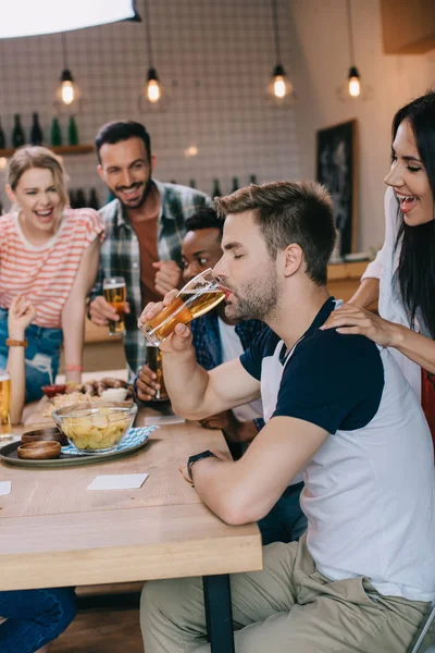 Young man drinking light beer with closed eyes while spending time in pub together with cheerful multicultural friends — Stock Photo