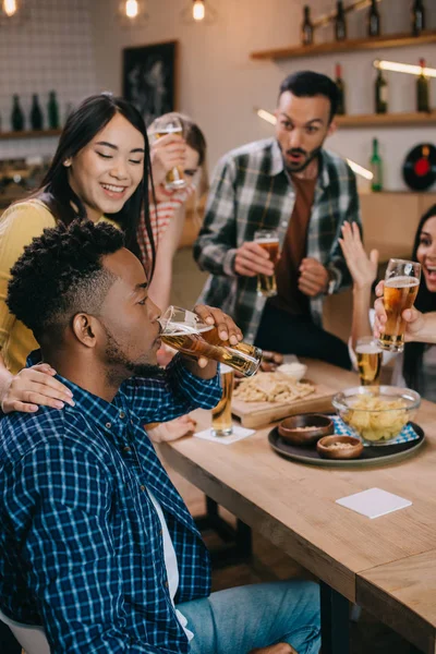 Messa a fuoco selettiva di bell'uomo afro-americano che beve birra leggera mentre trascorre del tempo in pub insieme a allegri amici multiculturali — Foto stock