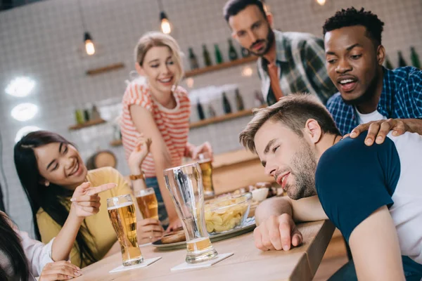 Cheerful multicultural friends looking at drunk young man in pub — Stock Photo