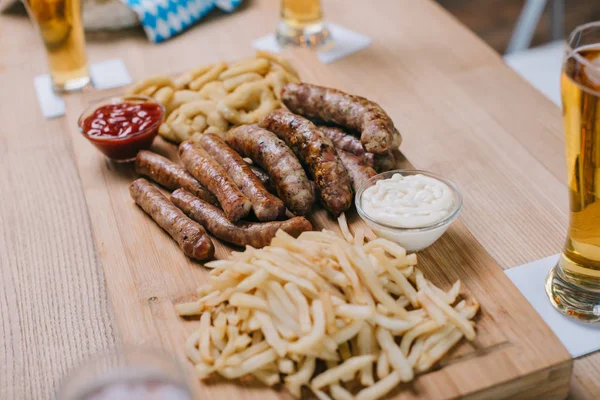 Vassoio di legno con salsicce fritte, anelli di cipolla, patatine fritte e salse vicino a bicchieri con birra leggera in pub — Foto stock