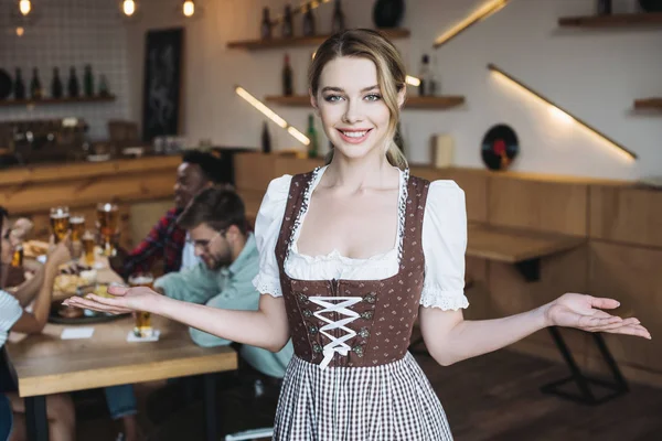 Hermosa camarera en traje nacional alemán mirando a la cámara y mostrando gesto de bienvenida - foto de stock