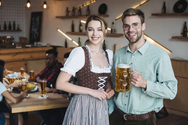 Hermosa camarera en traje nacional alemán de pie cerca de hombre joven sosteniendo taza de cerveza ligera y sonriendo a la cámara - foto de stock