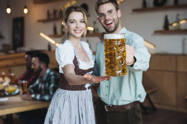 Enfoque selectivo de alegre joven sosteniendo taza de cerveza lager mientras está de pie cerca de hermosa camarera en traje alemán tradicional - foto de stock