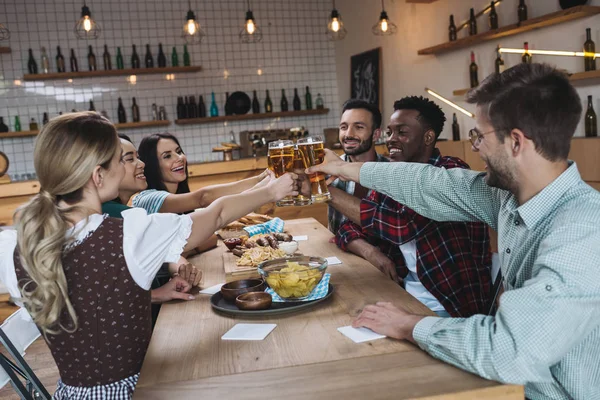 Amigos multiculturais alegres batendo copos de cerveja leve enquanto comemorava octoberfest no pub — Fotografia de Stock