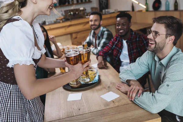 Vista recortada de la camarera sosteniendo vasos de cerveza ligera cerca de amigos multiculturales - foto de stock