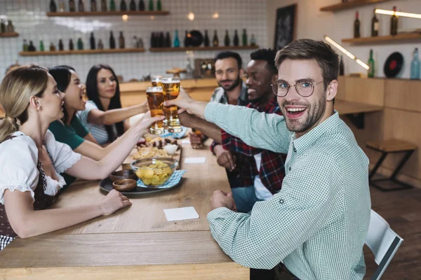Joven guapo sonriendo a la cámara mientras celebra octoberfest en el pub junto con amigos multiculturales - foto de stock