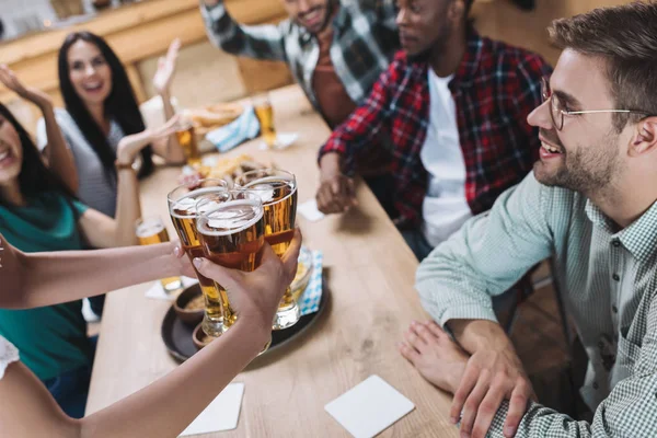 Vista parcial de la camarera sosteniendo vasos de cerveza ligera cerca de amigos multiculturales - foto de stock