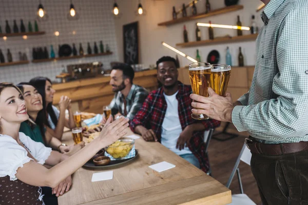 Vista cortada do homem segurando copos de cerveja leve perto de amigos multiculturais no pub — Stock Photo