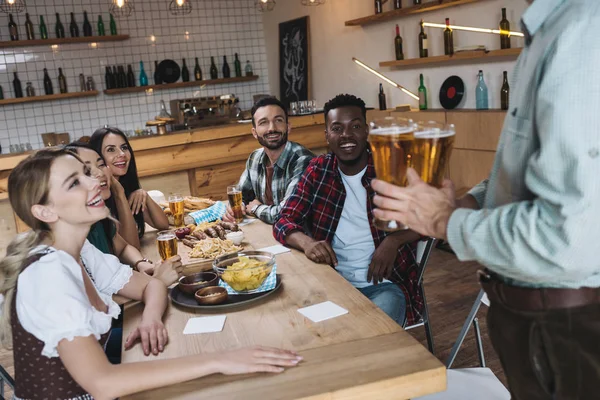 Vue partielle de l'homme tenant des verres de bière légère près d'amis multiculturels dans un pub — Photo de stock