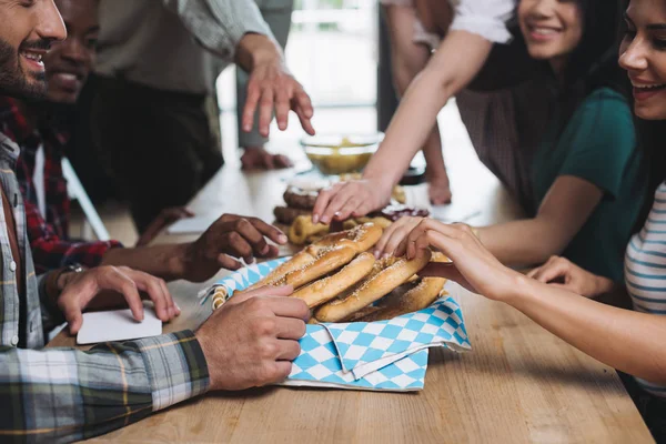 Vista recortada de amigos multiculturales tomando sabrosos pretzels mientras pasan tiempo juntos en el pub - foto de stock