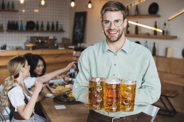 Jovem bonito segurando canecas com cerveja e sorrindo para a câmera — Fotografia de Stock