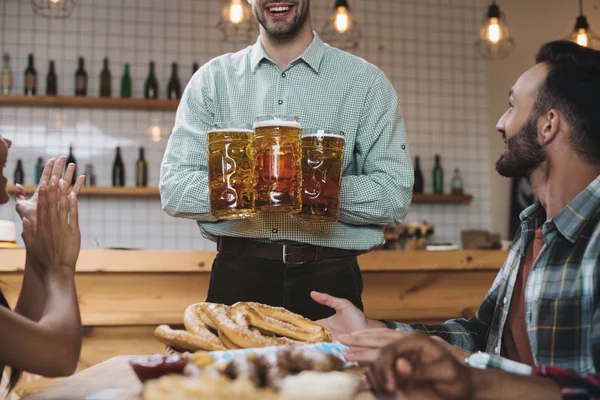 Vista recortada de un joven sosteniendo tazas con cerveza mientras está de pie cerca de amigos multiculturales en el pub - foto de stock