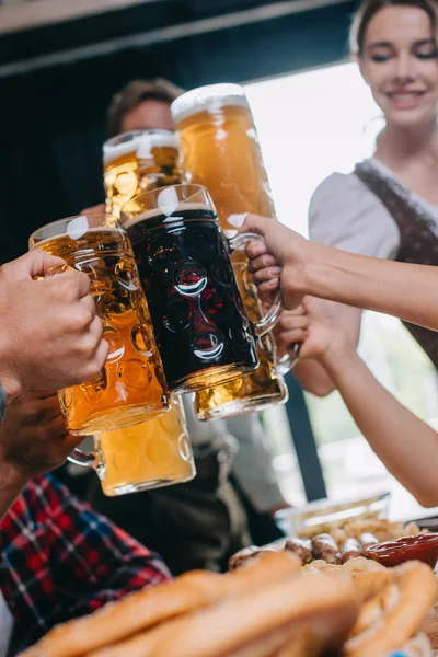 Partial view of friends clinking mugs of light and dark beer while celebrating octoberfest in pub — Stock Photo