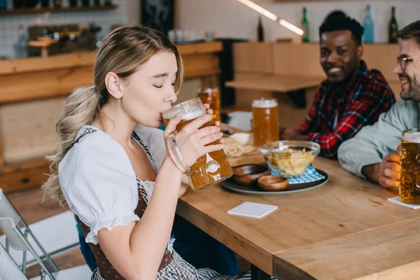 Selective focus of young woman in traditional german costume drinking beer near multicultural friends — Stock Photo