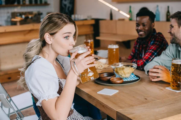 Foyer sélectif de la jeune femme en costume traditionnel allemand boire de la bière près de joyeux amis multiculturels — Photo de stock