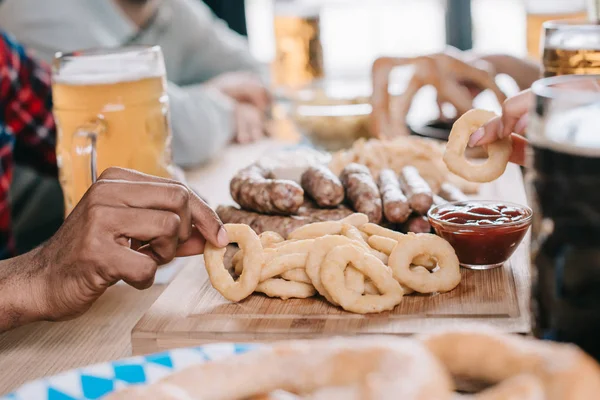 Cropped view of multicultural friends sitting at table with fried onion rings, sausages and mugs of beer in pub — Stock Photo