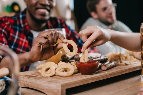 Vista ritagliata di amici multiculturali che mangiano anelli di cipolla fritti con ketchup in pub — Foto stock