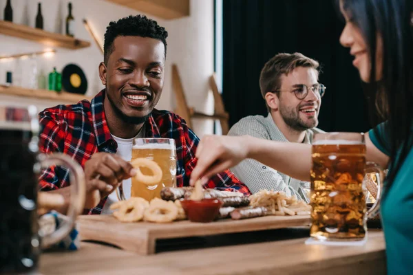 Amici multiculturali sorridenti che mangiano anelli di cipolla fritti con ketchup in pub — Foto stock