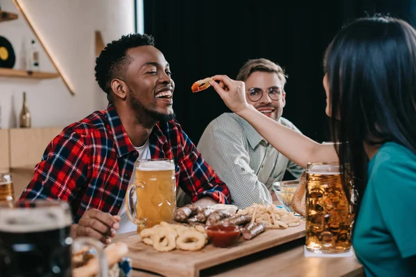 Rückansicht einer jungen Frau, die einen afrikanisch-amerikanischen Mann mit gebratenem Zwiebelring füttert, während sie in einer Kneipe Oktoberfest feiert — Stockfoto