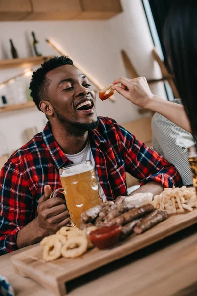 Vista recortada de la mujer joven que alimenta al hombre afroamericano con anillo de cebolla frita mientras celebra el octoberfest en el pub - foto de stock
