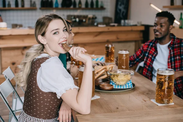 Fröhliche junge Frau isst Bratwurst, während sie mit multikulturellen Freunden Oktoberfest feiert — Stockfoto