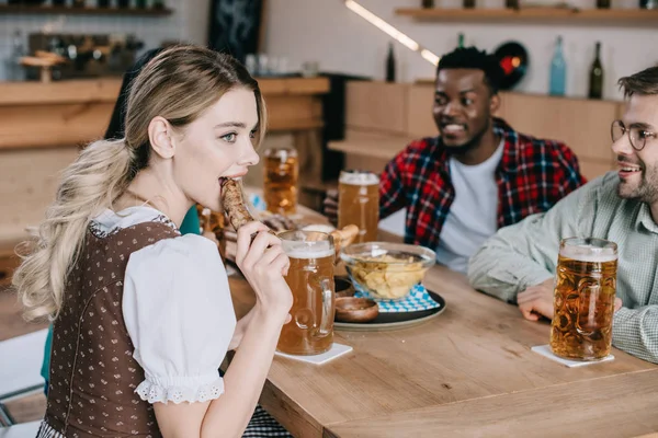 Junge Frau isst Bratwurst, während sie mit fröhlichen multikulturellen Freunden Oktoberfest feiert — Stockfoto