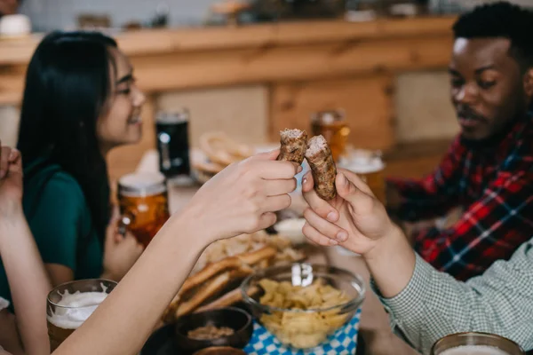 Vista ritagliata di giovane donna e donna che tiene salsiccia fritta mentre celebra l'octoberfest con amici multiculturali — Foto stock