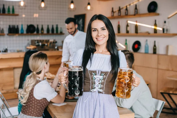 Hermosa camarera en traje alemán tradicional sosteniendo tazas de cerveza clara y oscura y sonriendo a la cámara - foto de stock