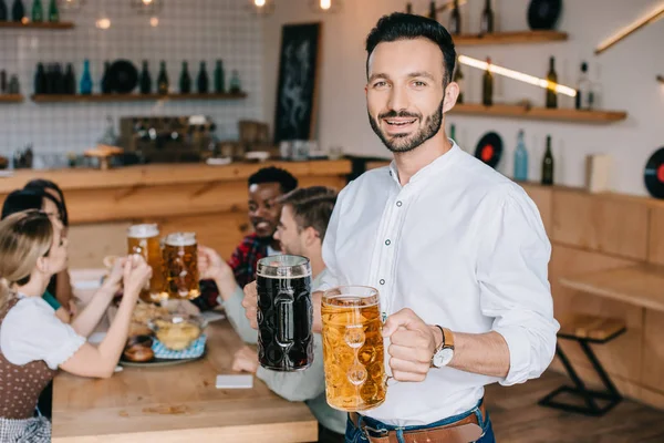 Joven guapo sosteniendo tazas de cerveza clara y oscura y sonriendo a la cámara - foto de stock