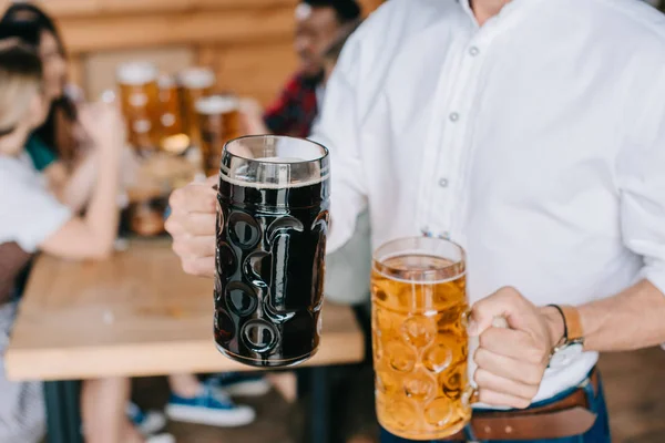 Vista recortada de un joven sosteniendo tazas de cerveza clara y oscura en el pub - foto de stock