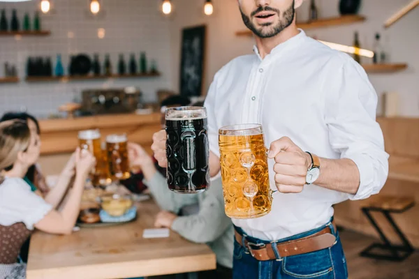 Vista parcial del joven sosteniendo tazas de cerveza clara y oscura en el pub — Stock Photo