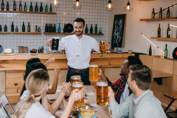 Alegre joven sosteniendo tazas de cerveza clara y oscura en el pub cerca de amigos multiculturales - foto de stock