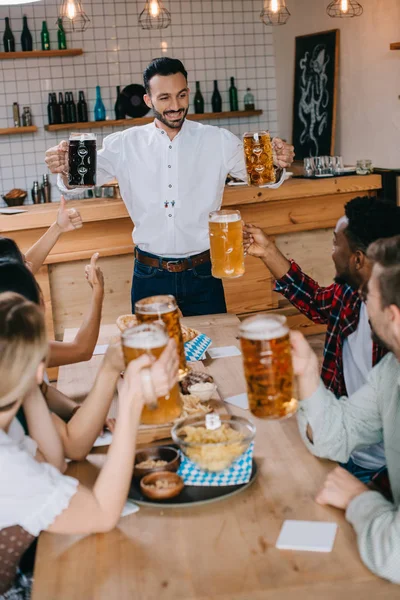 Joven guapo sosteniendo tazas de cerveza clara y oscura en el pub cerca de amigos multiculturales - foto de stock