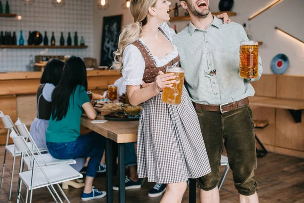 Cropped view of cheerful man and woman in traditional german costumes holding mugs of beer — Stock Photo