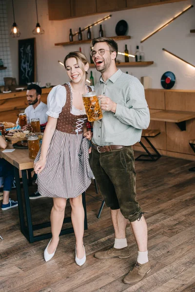 Jovem homem e mulher em trajes tradicionais alemães segurando canecas de cerveja — Fotografia de Stock