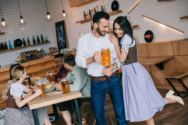 Jovem bonito segurando copo de cerveja enquanto estava perto de mulher atraente em traje alemão tradicional — Fotografia de Stock