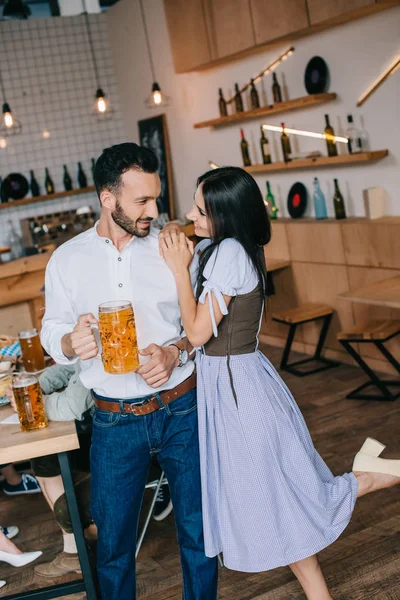 Smiling young man holding glass of beer while standing near beautiful woman in traditional german costume — Stock Photo