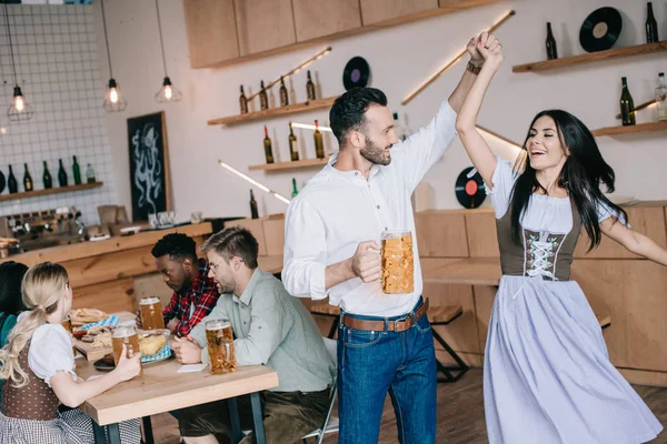 Handsome young man holding glass of beer while dancing with beautiful woman in traditional german costume — Stock Photo