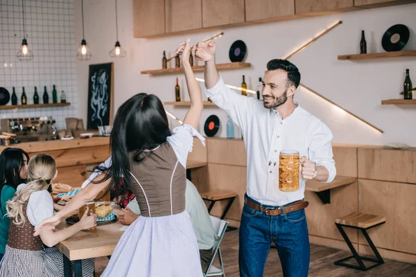 Jovem feliz segurando copo de cerveja enquanto dança com a jovem mulher em traje alemão tradicional — Fotografia de Stock