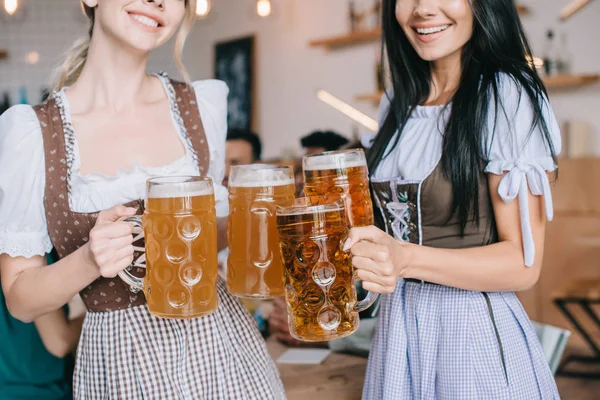 Обрезанный вид на двух молодых официанток в традиционных немецких костюмах, держащих кружки с пивом — стоковое фото