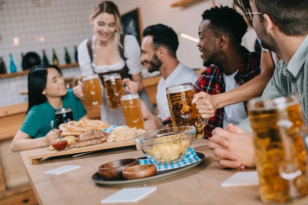 Selective focus of waitress in traditional german costume serving beer for multicultural friends in pub — Stock Photo