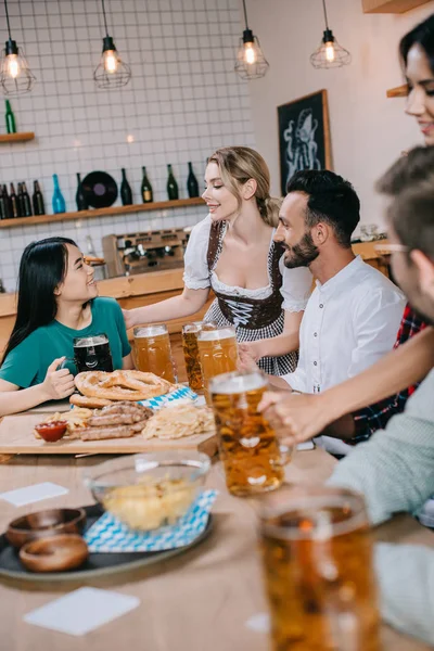 Two waitresses in traditional german costume serving beer for multicultural friends in pub — Stock Photo