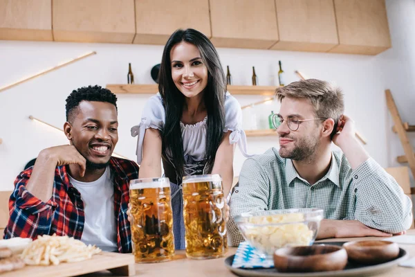 Atractiva camarera en traje tradicional alemán sirviendo cerveza para amigos multiculturales en el pub - foto de stock