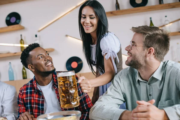 Beautiful waitress in traditional german costume serving beer for multicultural friends in pub — Stock Photo